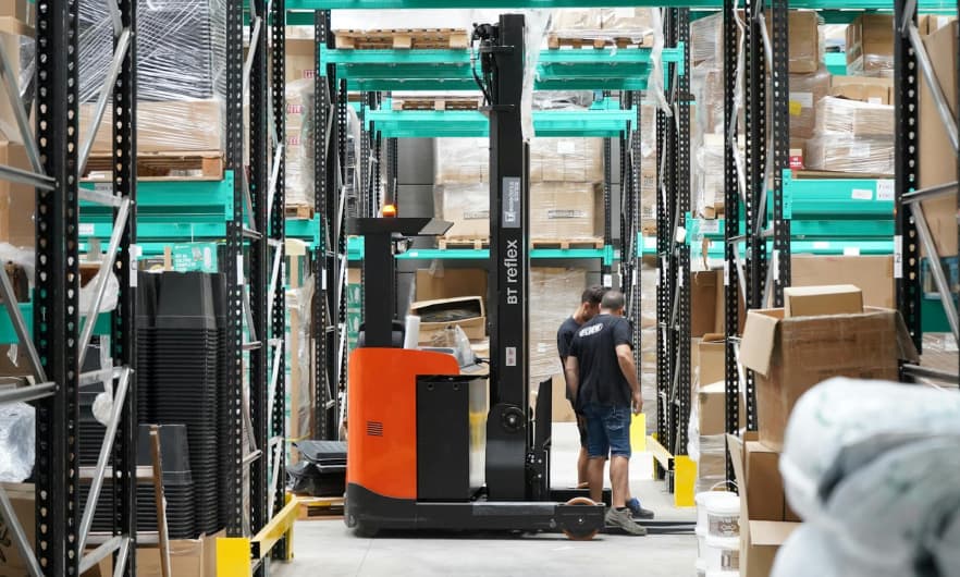 A worker operates a forklift in a warehouse aisle filled with shelves and boxes.